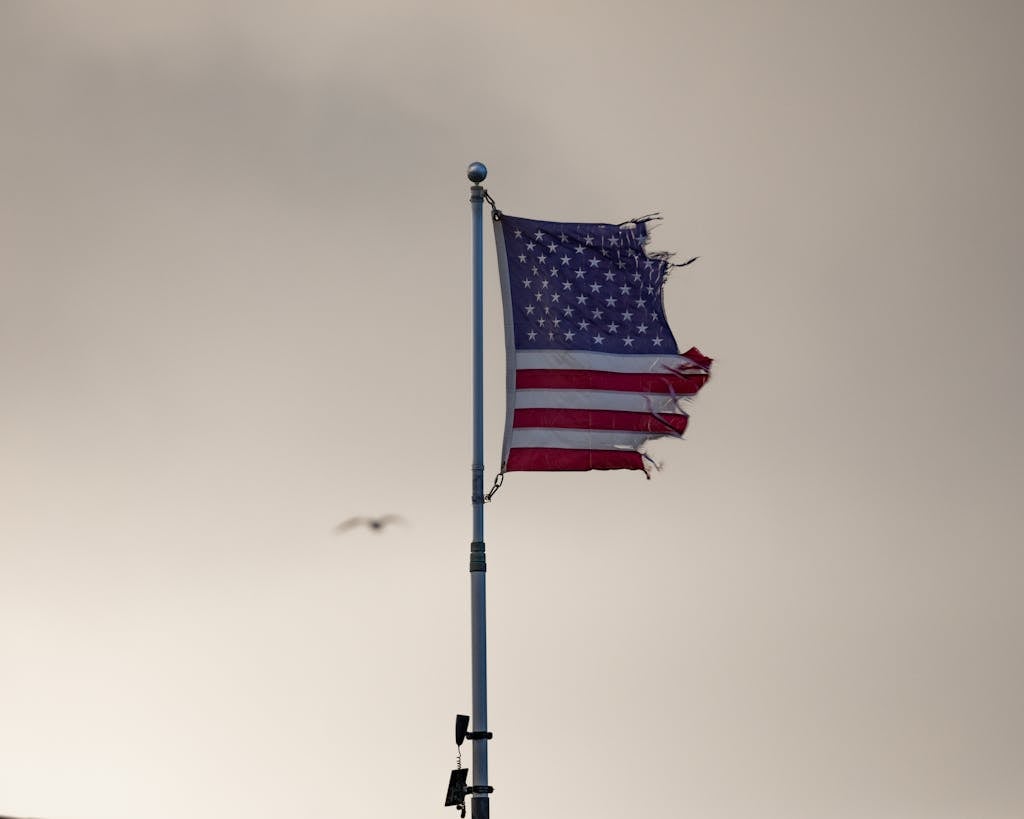 A tattered American flag waving on a flagpole with an overcast backdrop. Symbolic imagery.