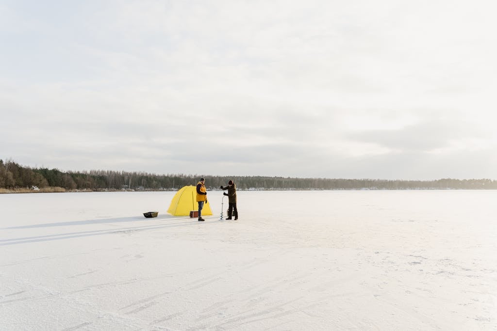 Fishermen ice fishing on a frozen lake with a yellow tent under a cloudy sky.